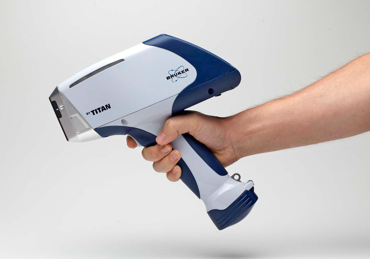 A hand holding a blue and white handheld device labeled "Bruker S1 TITAN" against a plain, light background. The device has a sleek, ergonomic design and appears to be used for scanning or analyzing materials.